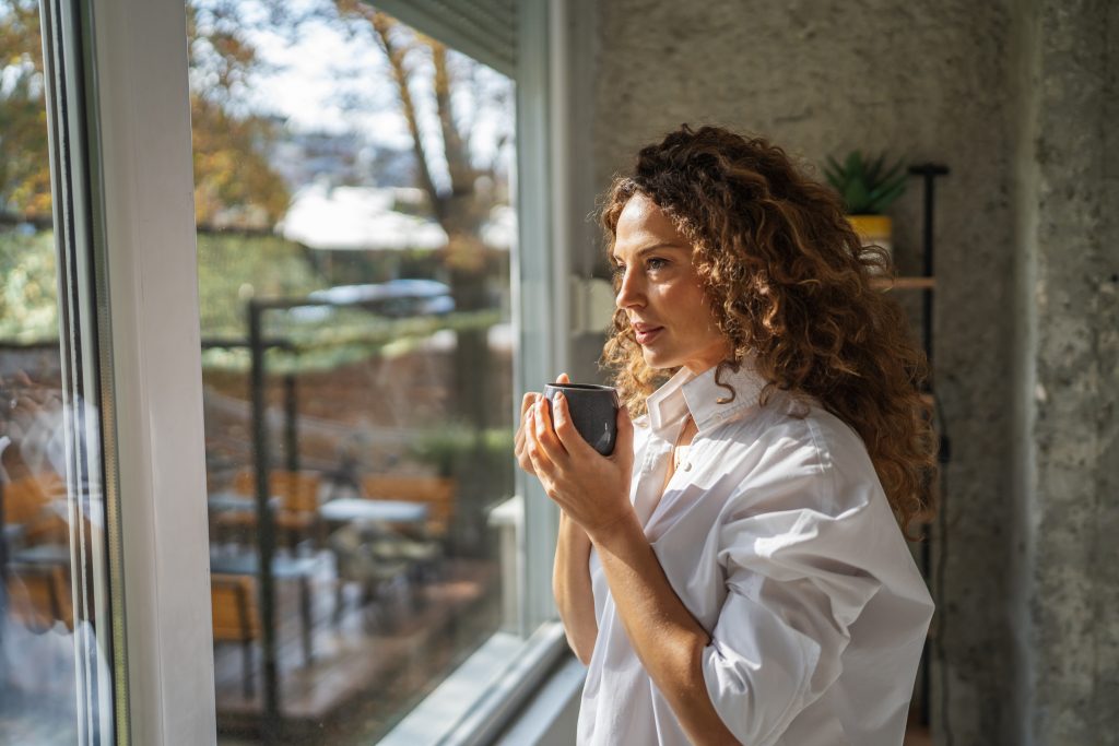 woman with curly brown hair in a white shirt smiles while holding a mug, looking through a window