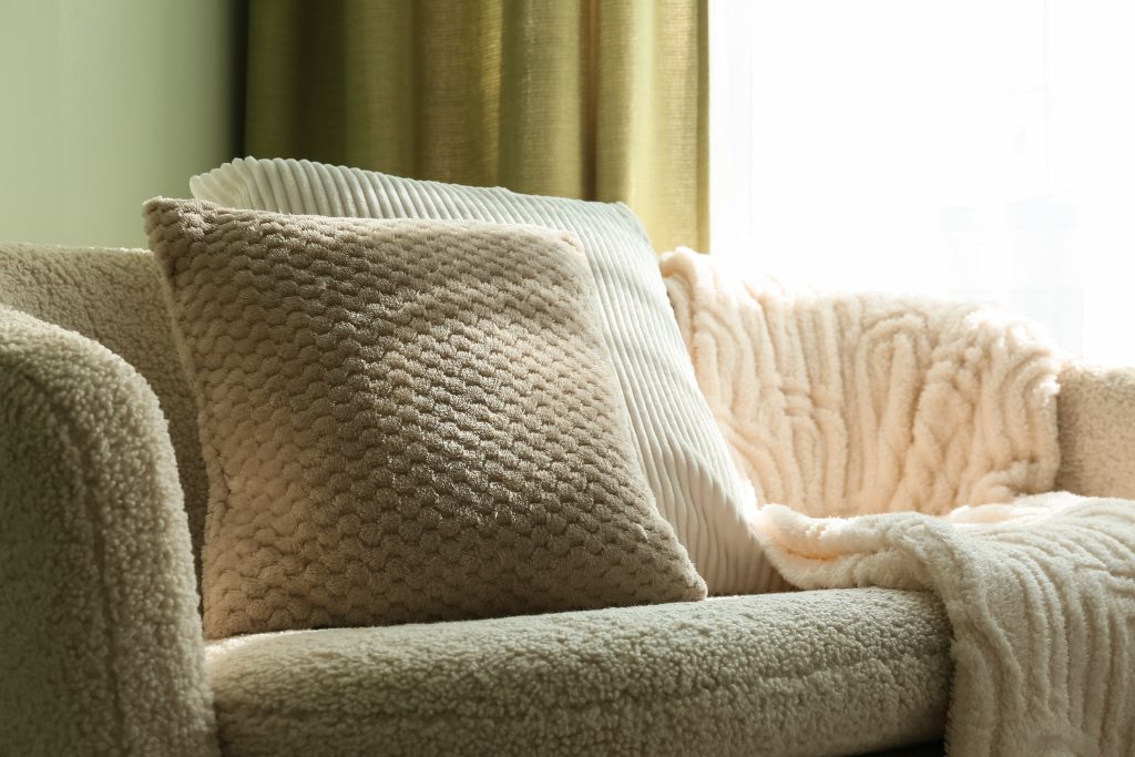 Cozy home interior with soft textured pillows and blanket on a comfortable sofa by a window with warm sunlight, warm sunlight streams through a nearby window